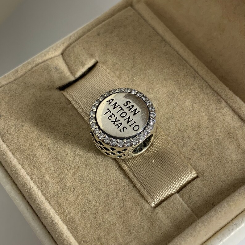 Puede incluir: Encanto de plata con un borde redondo, blanco y brillante y las palabras "SAN ANTONIO TEXAS" en letras negras.