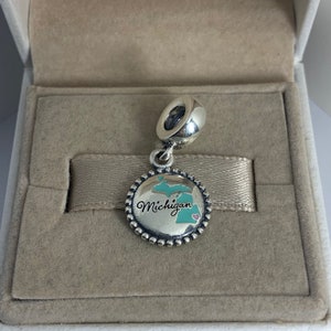 Puede incluir: Un charm de plata con un contorno de esmalte azul y verde del estado de Michigan. El charm tiene un borde de cuentas de plata y la palabra "Michigan" escrita en cursiva en el charm. Un pequeño corazón rosa se encuentra en la esquina inferior derecha del charm.