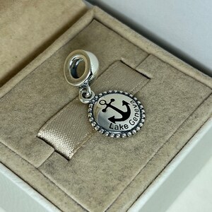 Puede incluir: Encanto de plata con un ancla negra y el texto "Lake Geneva" en un disco redondo.