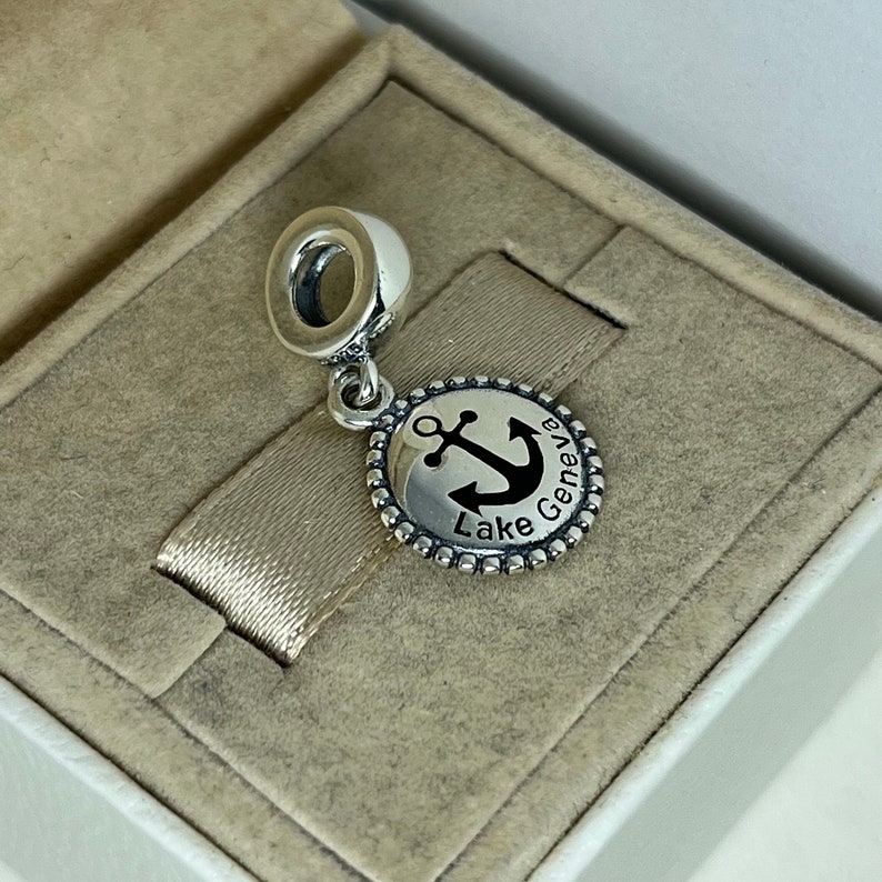 Puede incluir: Encanto de plata con un ancla negra y el texto "Lake Geneva" en un disco redondo.