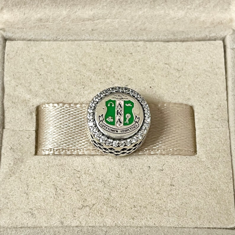 May include: A silver charm with a green and white enamel shield featuring the letters "AKKA" and the words "Great Women" and "For Times To Come". The charm is surrounded by a circle of clear cubic zirconia stones.
