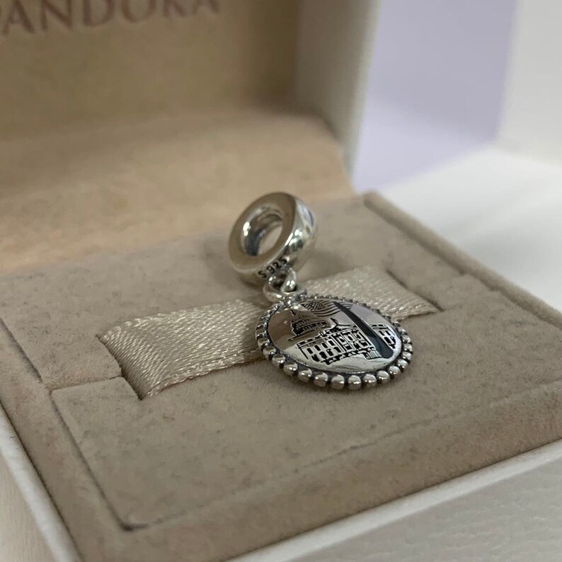 Puede incluir: Un charm de plata con una imagen detallada de un edificio, posiblemente un edificio gubernamental, con una peque&ntilde;a bandera estadounidense en la parte superior. El charm est&aacute; en una peque&ntilde;a caja blanca con un interior marr&oacute;n.