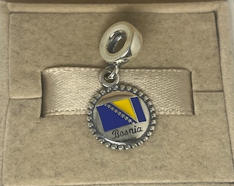 Könnte beinhalten: Silberner Anhänger mit einer blau-gelben Emaille-Flagge von Bosnien. Der Anhänger hat einen perlenbesetzten Rand und das Wort "Bosnien" steht unter der Flagge.