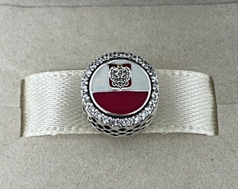 May include: A silver charm with a white and red enamel design featuring the Polish flag and coat of arms. The charm is surrounded by clear cubic zirconia stones.