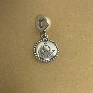 May include: Silver charm with a round, beaded edge. The charm features a silver circle with a crown and the word "PANDORA" inscribed around it.