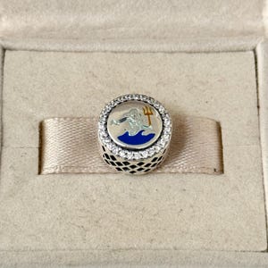 May include: A silver charm with a blue and gold enamel design of a mythical sea creature holding a trident. The charm is surrounded by a circle of clear cubic zirconia stones.
