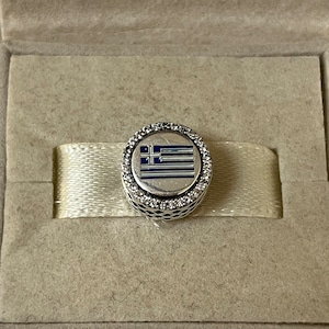 May include: A silver charm with a blue and white enamel Greek flag design. The charm is surrounded by a circle of clear cubic zirconia stones.