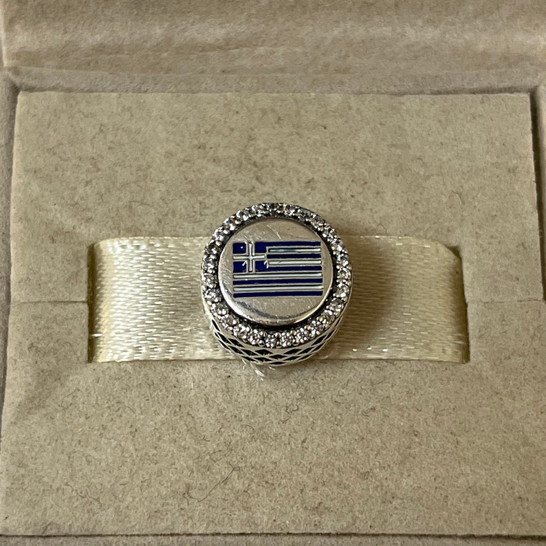 May include: A silver charm with a blue and white enamel Greek flag design. The charm is surrounded by a circle of clear cubic zirconia stones.