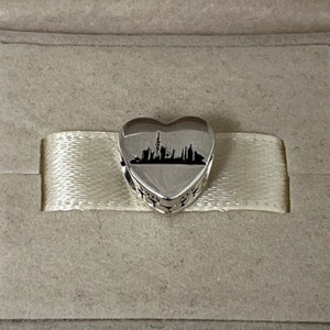 Może przedstawiać: Srebrny charm w kształcie serca z czarną sylwetką panoramy miasta, w tym wysokiego budynku ze spiczastym dachem.