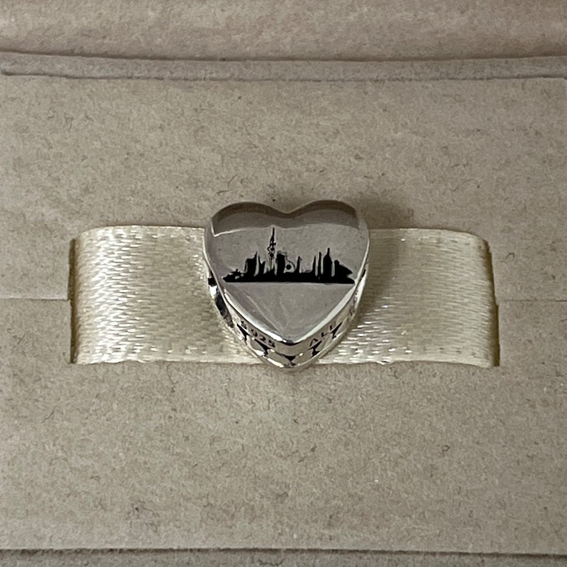 Może przedstawiać: Srebrny charm w kształcie serca z czarną sylwetką panoramy miasta, w tym wysokiego budynku ze spiczastym dachem.
