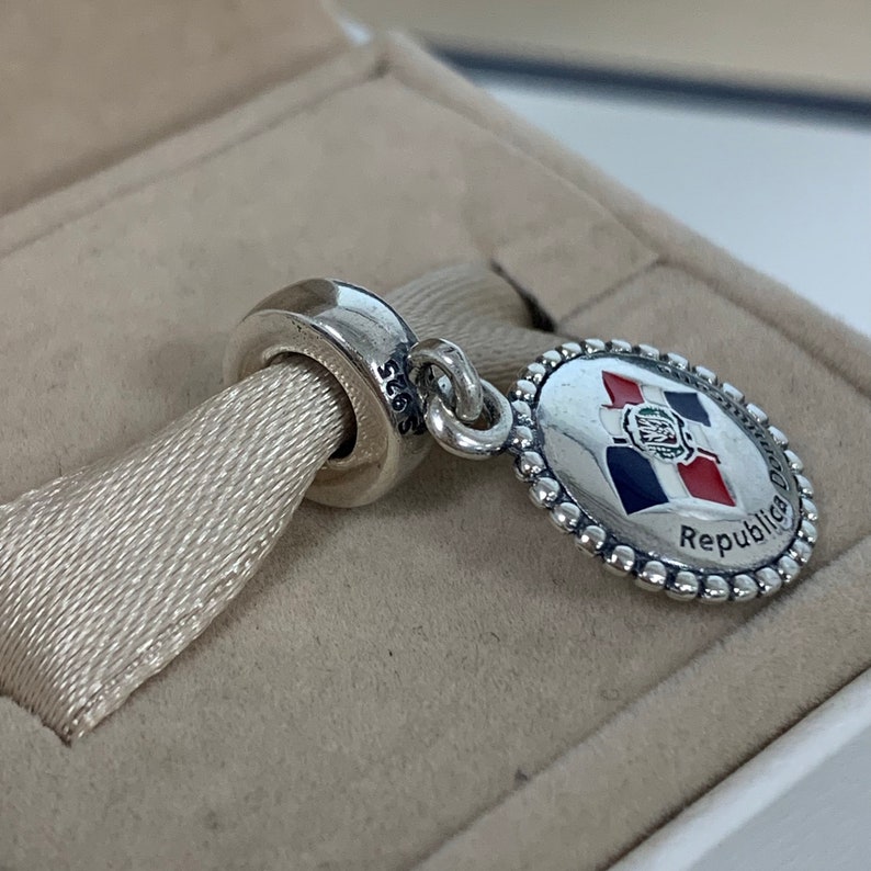 May include: Silver charm featuring a round, beaded border and a blue, white, and red enamel design of the Dominican Republic flag. The charm has the text "Republica Dominicana" on the bottom and "S925" on the back.