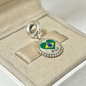 May include: Silver charm with a green, yellow, and blue heart-shaped charm with the word "Brazil" on it.