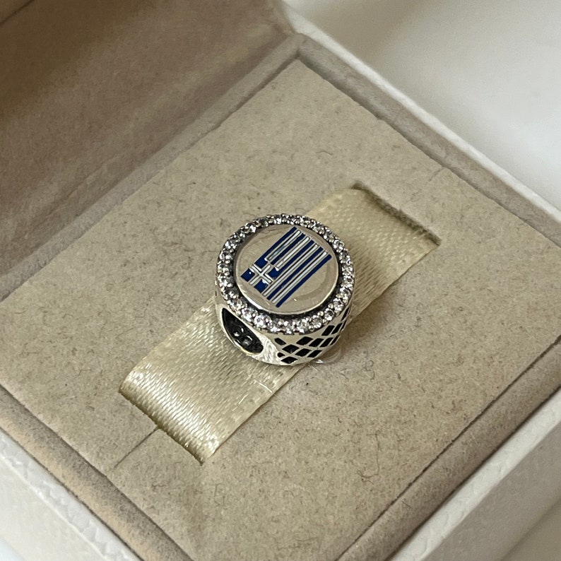 May include: A silver charm with a blue and white enamel Greek flag design. The charm is surrounded by a circle of clear cubic zirconia stones.