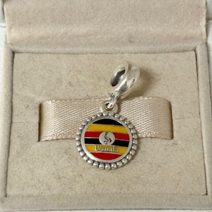 May include: A silver charm with a round pendant featuring the flag of Uganda. The charm has a beaded edge and the word "Uganda" in red. The flag design includes black, yellow, and red stripes, with a crane emblem.