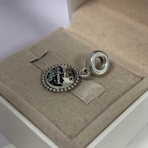 May include: Silver charm with a round, black and white enamel design featuring the word "Hollywood" and a palm tree silhouette. The charm has a silver bead border and a silver loop for attaching to a bracelet.
