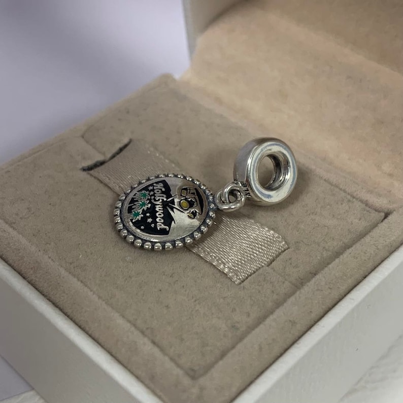May include: Silver charm with a round, black and white enamel design featuring the word "Hollywood" and a palm tree silhouette. The charm has a silver bead border and a silver loop for attaching to a bracelet.