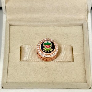 Peut inclure: Un charm en or rose avec un motif de grenouille verte sur fond noir, entouré de cristaux transparents. Le charm est présenté dans un écrin beige avec un ruban de satin.