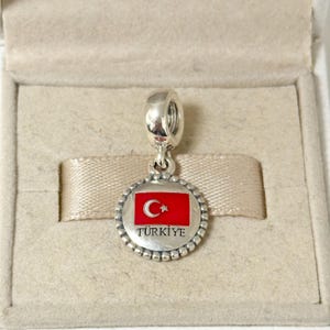 Puede incluir: Un charm de plata con una bandera turca de esmalte rojo y la palabra "TÜRKİYE" en letras negras.