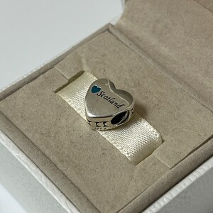 May include: A silver charm in the shape of a heart with the word "Scotland" inscribed on it. The heart has two small blue hearts on the top. The charm is in a white box.
