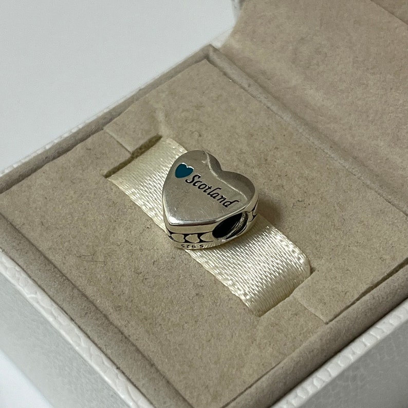 May include: A silver charm in the shape of a heart with the word "Scotland" inscribed on it. The heart has two small blue hearts on the top. The charm is in a white box.