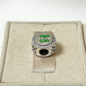 May include: Silver charm with a green and white enamel design featuring a crest with the letters "AKA" and the words "Founded 1908" around the crest. The charm is surrounded by a circle of clear cubic zirconia stones.