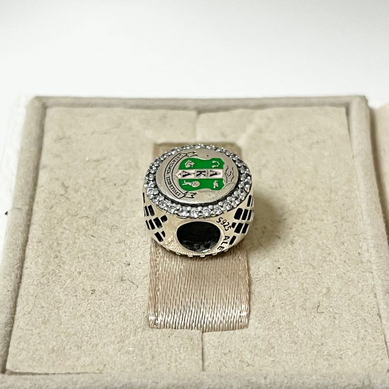 May include: Silver charm with a green and white enamel design featuring a crest with the letters "AKA" and the words "Founded 1908" around the crest. The charm is surrounded by a circle of clear cubic zirconia stones.