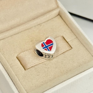 Puede incluir: Un dije de plata en forma de corazón con una incrustación de esmalte de la bandera de Noruega en colores rojo, blanco y azul. El lateral del dije tiene grabado el texto S 925, lo que indica una composición de plata de ley.