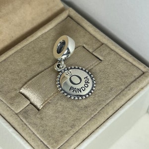 May include: A silver charm with a round disc featuring the Pandora logo and a crown above the letter "O".