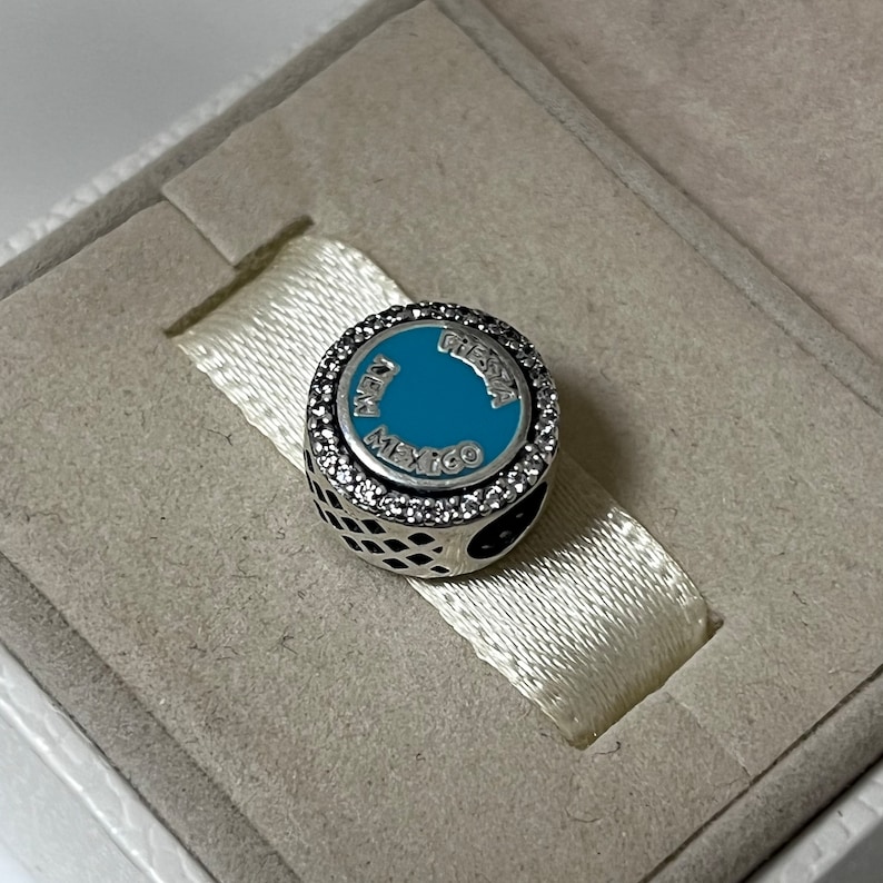 May include: A silver charm with a blue enamel circle featuring the words "Avey Mexico Fiesta" surrounded by clear cubic zirconia stones.