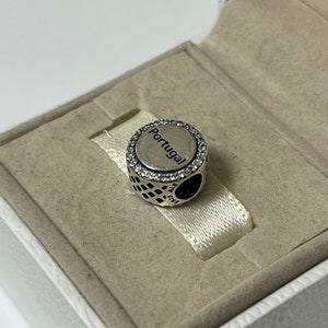 Puede incluir: Encanto de plata con una cara redonda y blanca que dice "Portugal" en letras negras. El encanto est&aacute; rodeado por un c&iacute;rculo de piedras de circonita c&uacute;bica transparentes. El encanto est&aacute; sentado en una caja blanca.