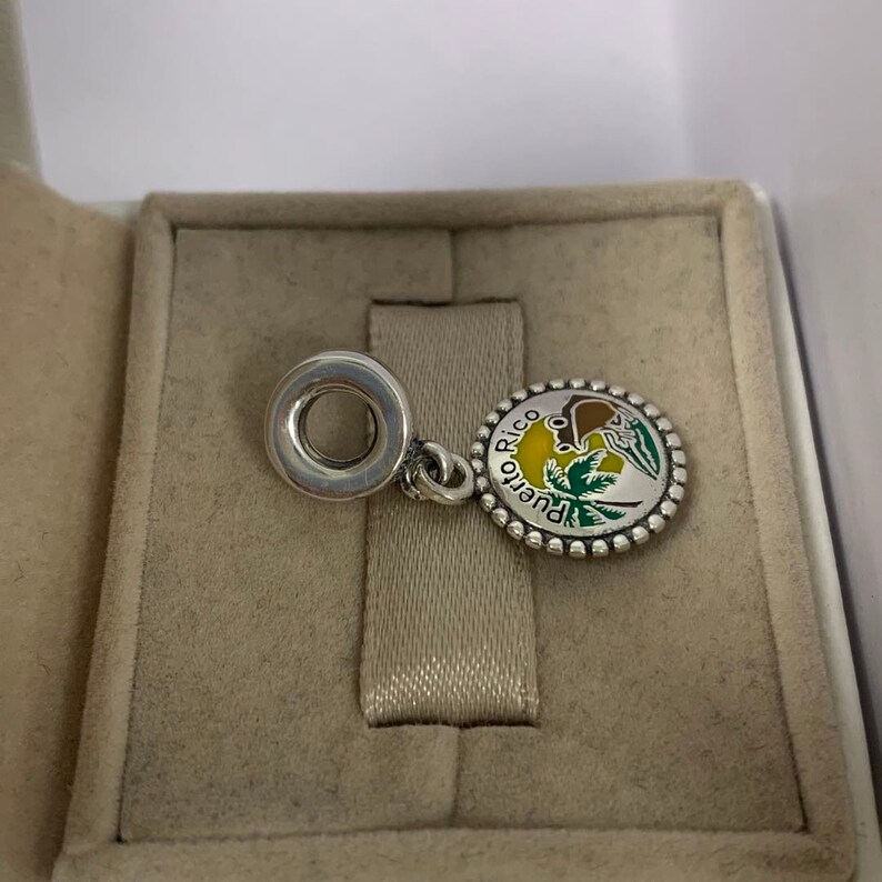 May include: A silver charm with a round pendant featuring a colourful design of Puerto Rico. The pendant has a yellow, green, and brown design with the words "Puerto Rico" around the edge. The charm is in a grey box.