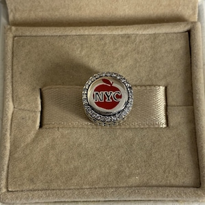 Könnte beinhalten: Ein silberner Anhänger mit einem roten Apfel-Design und dem Text "NYC" in Weiß. Der Anhänger ist von einer Reihe klarer Kristalle umgeben.