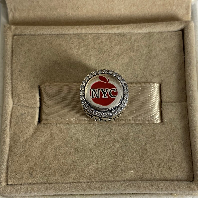 May include: A silver charm featuring a red apple design with the text "NYC" in white. The charm is encircled by a row of clear crystals.