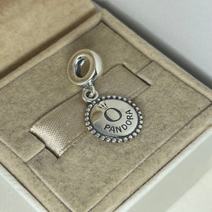 Puede incluir: Encanto de plata con un disco redondo de plata que presenta la palabra "Pandora" y una corona sobre la letra "O".