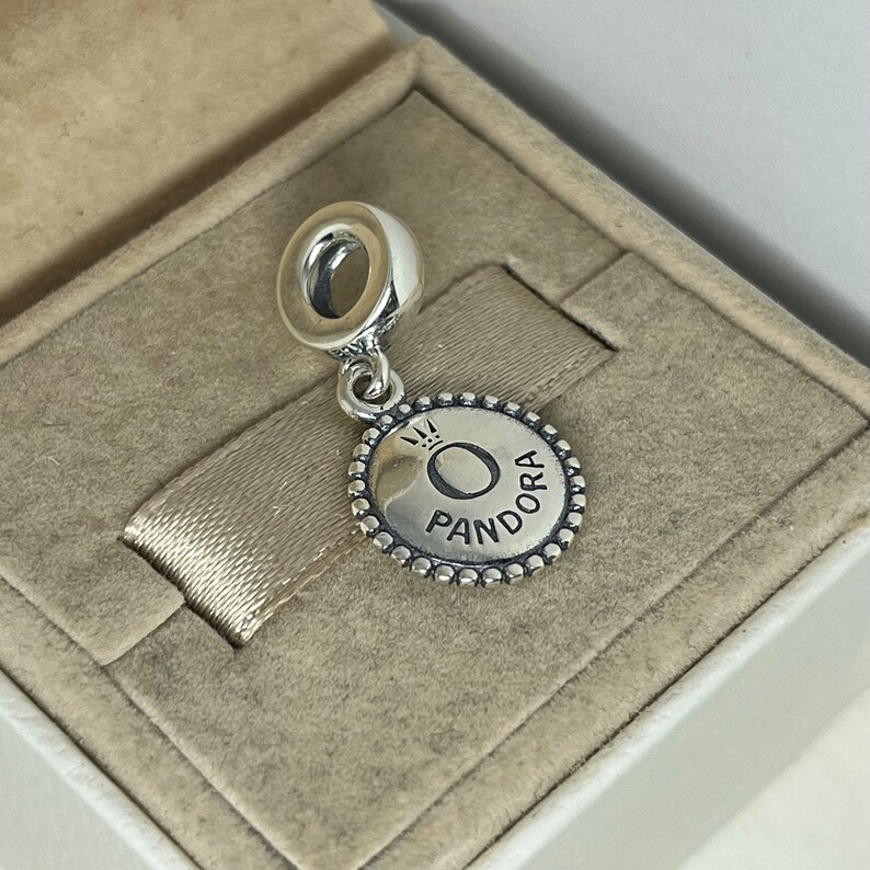 Puede incluir: Encanto de plata con un disco redondo de plata que presenta la palabra "Pandora" y una corona sobre la letra "O".