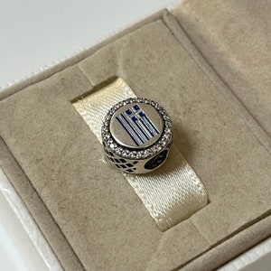 May include: A silver charm with a blue and white enamel Greek flag design surrounded by clear cubic zirconia stones.