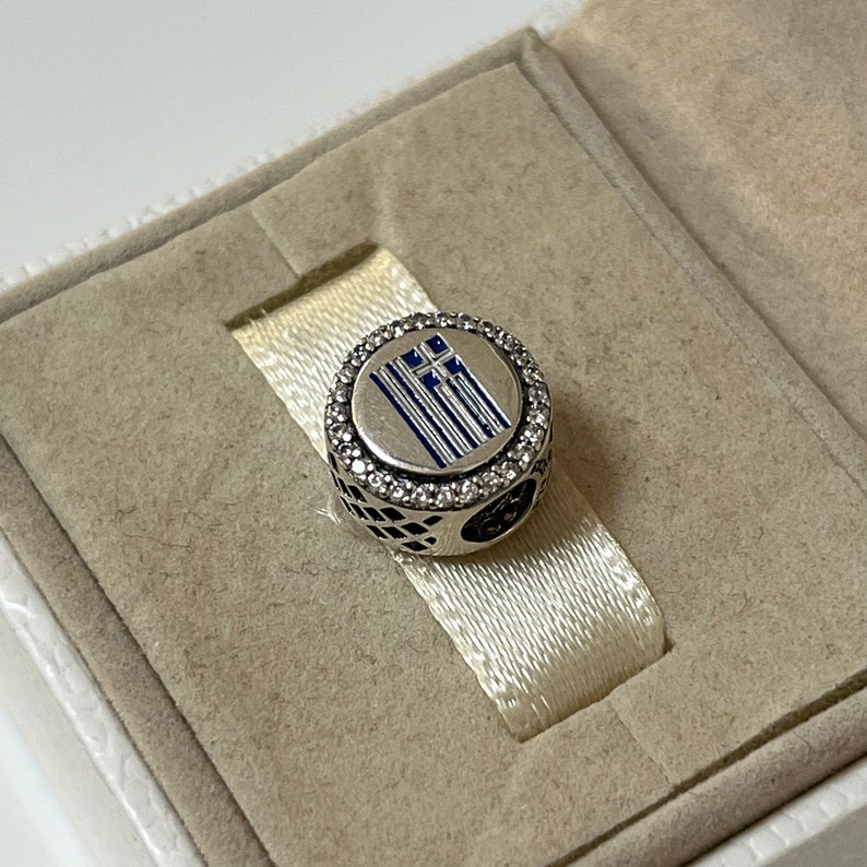 May include: A silver charm with a blue and white enamel Greek flag design surrounded by clear cubic zirconia stones.