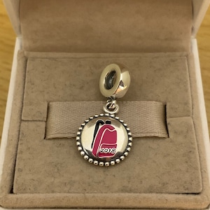 May include: A silver charm with a pink backpack design and the year 2018 printed on it.