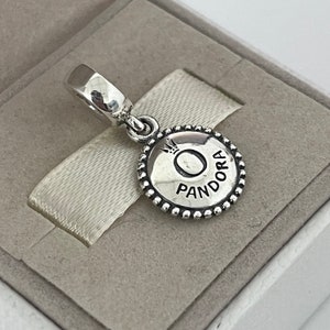 Op de afbeelding: Zilveren bedel met een ronde, parelrand. De bedel heeft een zilveren cirkel met het woord "Pandora" en een kroon boven de letter "O".