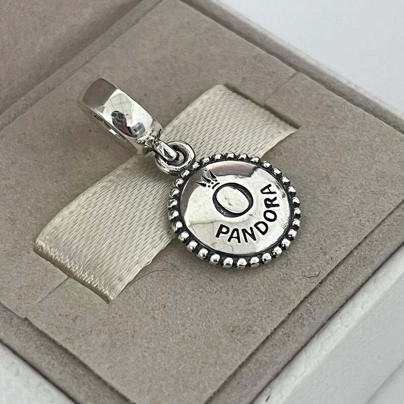 Op de afbeelding: Zilveren bedel met een ronde, parelrand. De bedel heeft een zilveren cirkel met het woord "Pandora" en een kroon boven de letter "O".