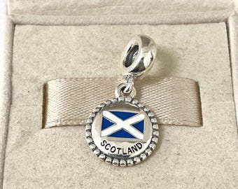 May include: A silver charm with a blue and white enamel Scottish flag design. The charm is round and has a beaded border. The word "SCOTLAND" is written in black lettering around the flag.