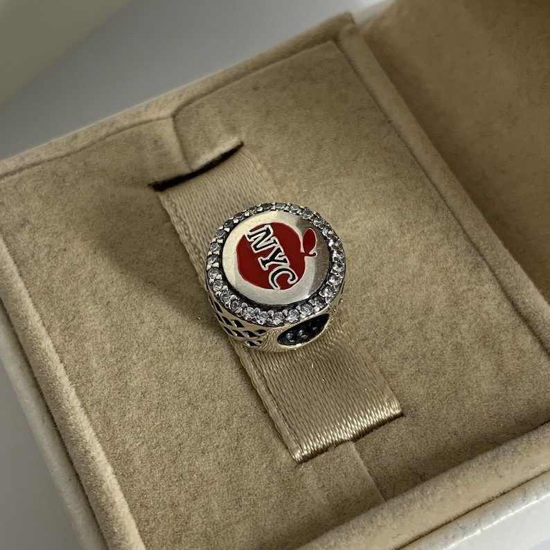 May include: A silver charm featuring a red apple design with the text "NYC" in white. The charm is surrounded by clear cubic zirconia stones.