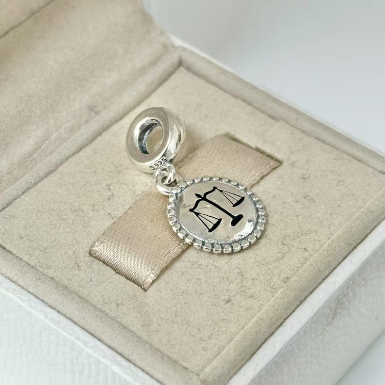 May include: A silver charm with a round pendant featuring a black scale of justice symbol. The charm is in a white box with a beige interior. The pendant has a beaded border and a loop for attaching to a bracelet or necklace.
