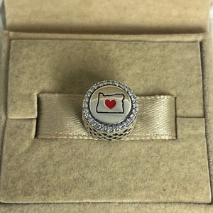 Peut inclure: Un charme en argent avec un contour blanc de l'État de l'Oregon et un cœur rouge au centre. Le charme est entouré d'un cercle de pierres de zircone cubique transparentes.