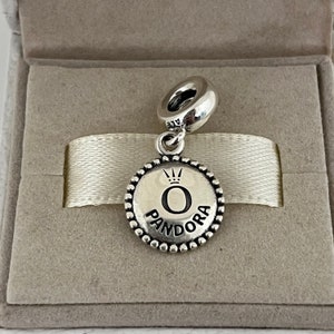 Op de afbeelding: Zilveren bedel met een ronde, parelrand. De bedel heeft een zwarte 'O' met een kroon erboven en het woord 'PANDORA' eronder.