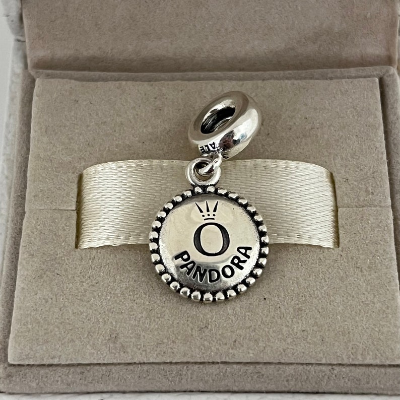 Op de afbeelding: Zilveren bedel met een ronde, parelrand. De bedel heeft een zwarte 'O' met een kroon erboven en het woord 'PANDORA' eronder.