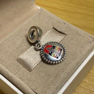May include: Silver charm with a red, yellow, and black enamel design. The charm features the Porsche logo and the text "20 Jahre 911 Carrera RS 2.7." The charm is in a white box with a beige interior.