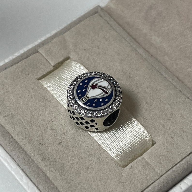 May include: A silver charm with a blue and white enamel hot air balloon design. The balloon has a red star and white stripes. The charm is surrounded by clear cubic zirconia stones.