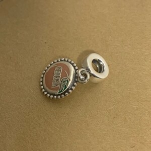 May include: Silver charm with a peach design and the word "GEORGIA" in black lettering. The charm has a small loop for attaching to a bracelet or necklace.