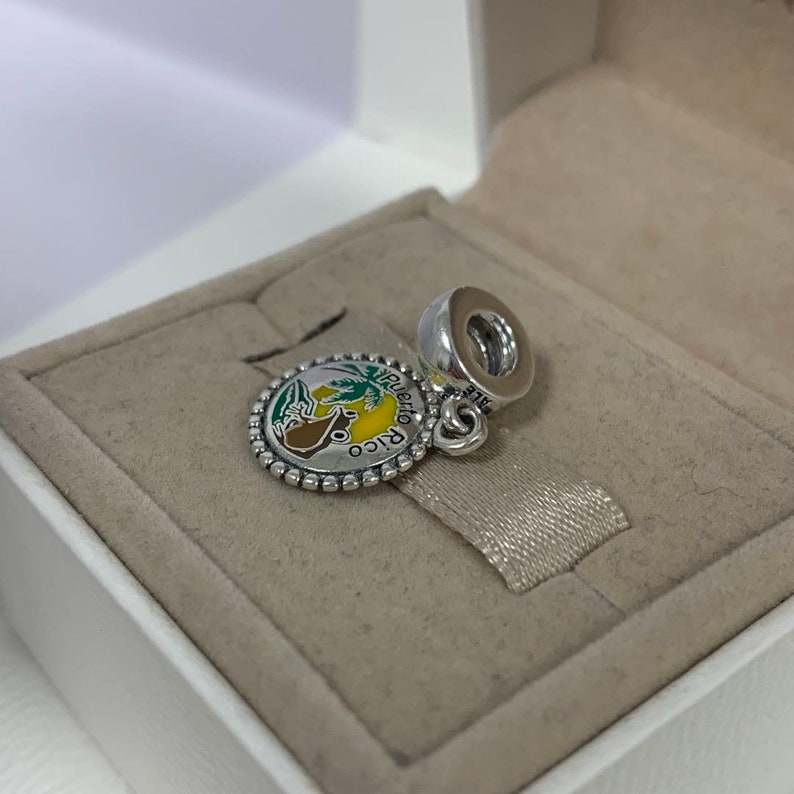 May include: A silver charm with a round, colourful design featuring a tropical scene with the words "Puerto Rico". The charm has a beaded edge and a loop for attaching to a bracelet or necklace.